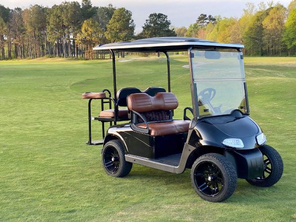 A black golf cart with brown seats is parked on a grassy golf course with trees in the background.