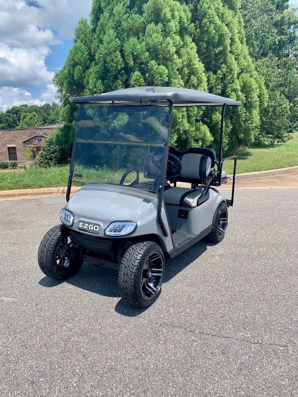 A gray E-Z-GO golf cart with a roof and windshield is parked on an asphalt surface, with trees and a house in the background.