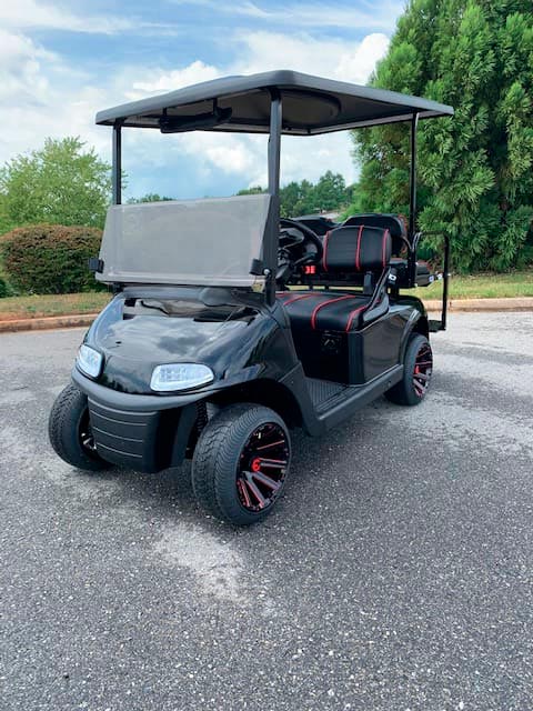 A black golf cart with red-trimmed seats is parked on an asphalt surface, surrounded by trees and bushes.