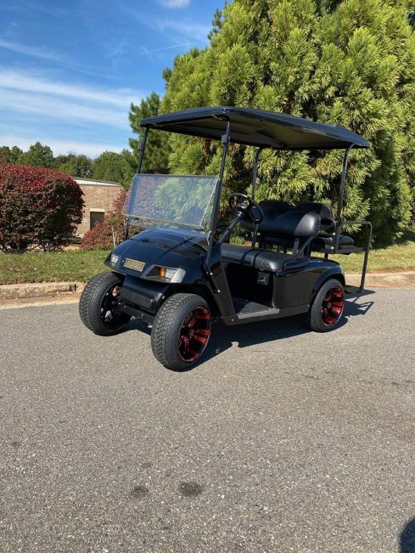 A black golf cart with red-trimmed wheels is parked on a paved road near green bushes and trees, with a brick building visible in the background.