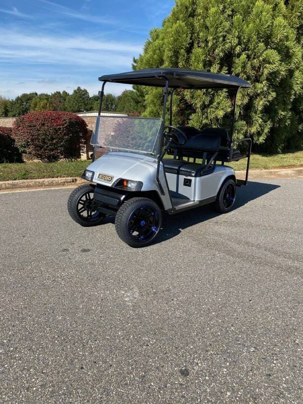 A silver golf cart with a black roof and seats is parked on a paved road beside green bushes and trees under a clear sky.