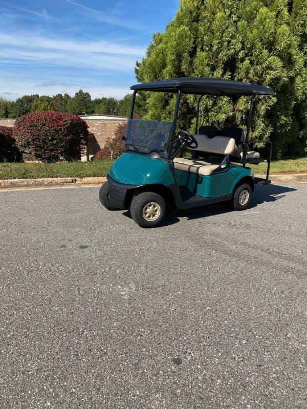 A green and black golf cart is parked on a paved road, with trees, bushes, and a low brick wall in the background under a clear sky.