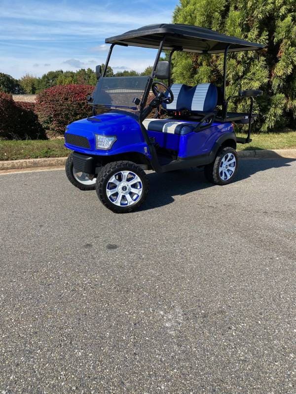 A blue golf cart with striped blue and white seats is parked on a paved surface, with bushes and trees in the background.