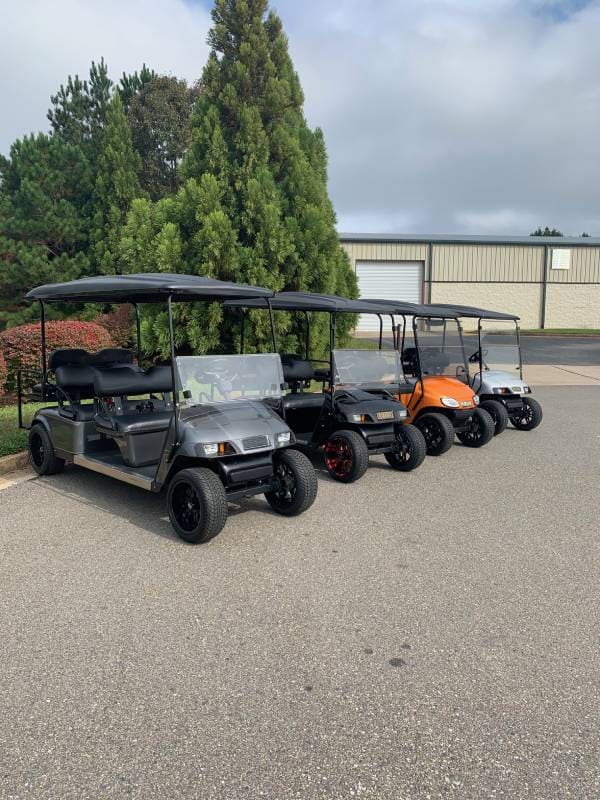 Four golf carts, including one orange, are parked in a row on a paved lot near trees and an industrial building.