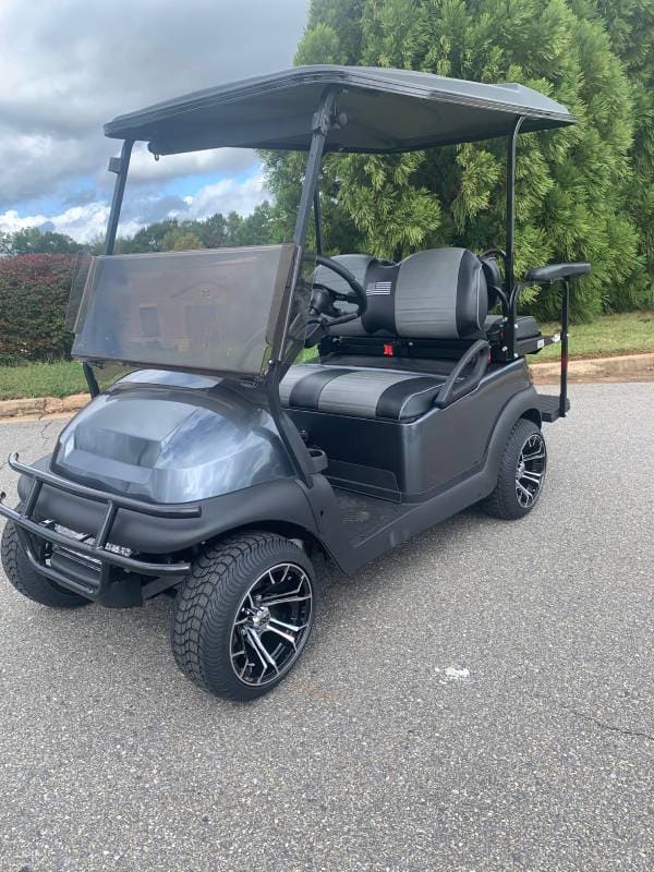 A black, two-seater golf cart with a roof and custom wheels is parked on a paved surface near greenery.