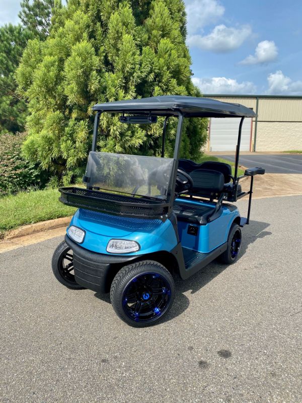 A blue golf cart with black roof and rims is parked on a paved road near greenery and a warehouse building under a partly cloudy sky.