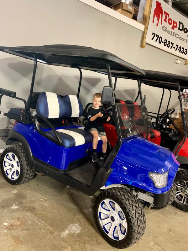 A young boy sits in the driver’s seat of a blue golf cart with black and white seats, parked indoors near other golf carts.