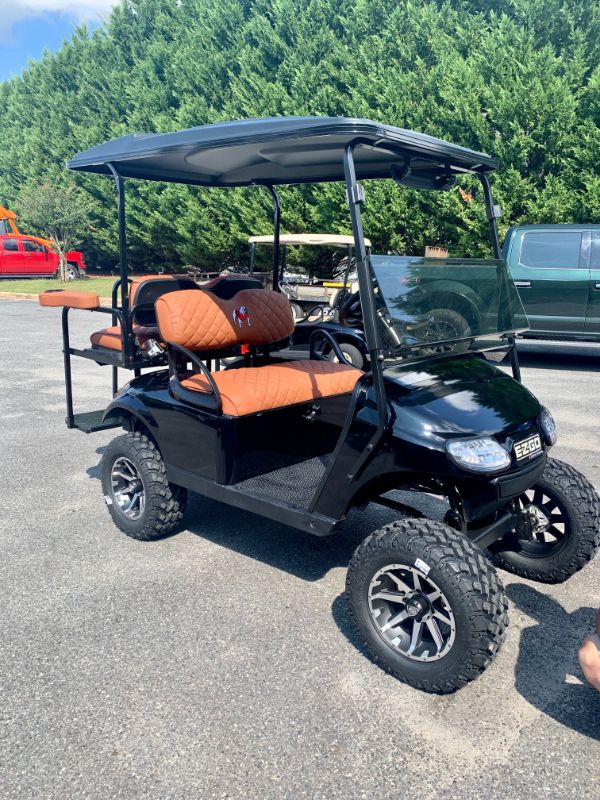 A black EZGO golf cart with brown diamond-stitched seats is parked on asphalt near other vehicles and green trees.