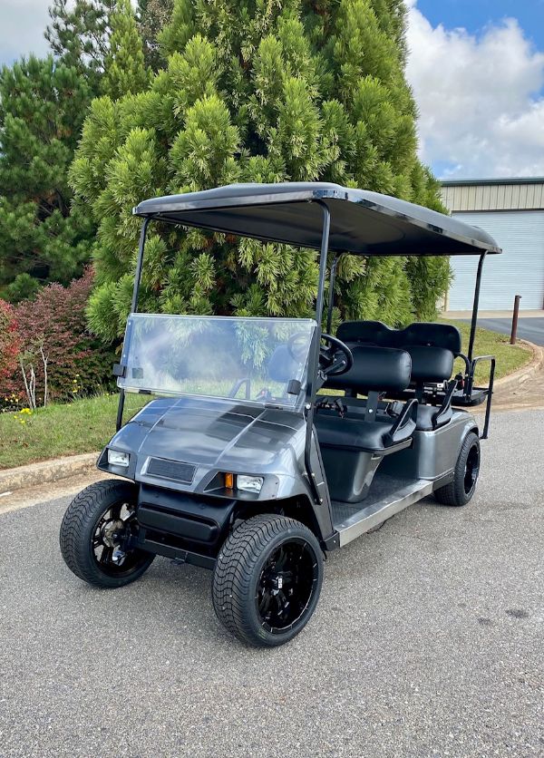 A black, four-seater golf cart is parked on a paved road near green trees and a building in the background.