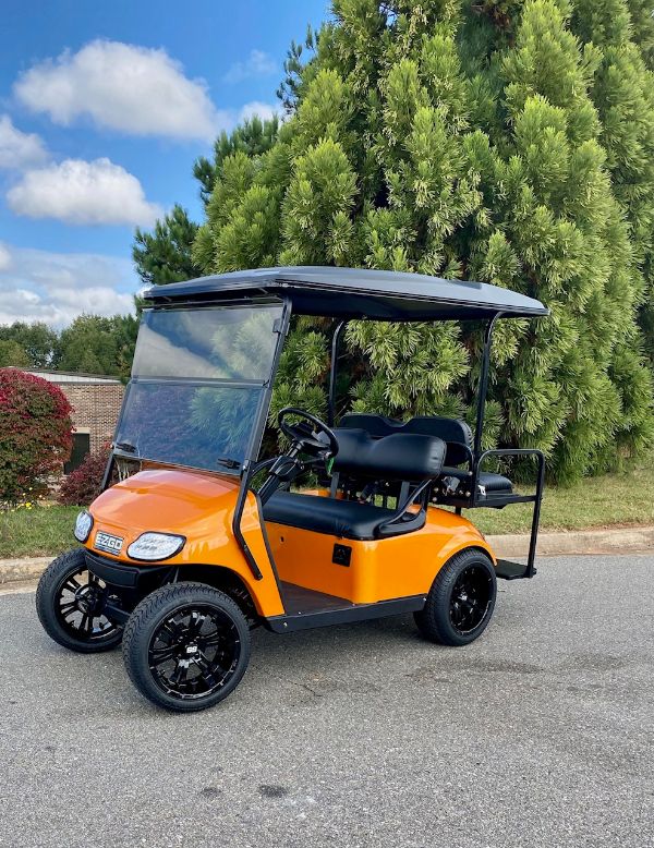 Bright orange golf cart with a black roof and black seats parked on a paved area near tall green trees under a partly cloudy sky.