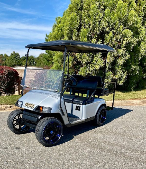 A silver golf cart with a black roof is parked on a paved road near green trees and shrubs under a blue sky.