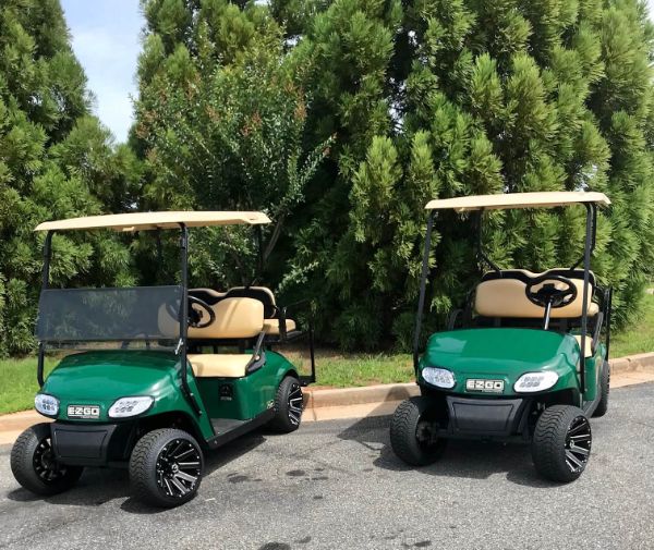 Two green E-Z-GO golf carts with beige seats are parked on a paved area near tall green trees.