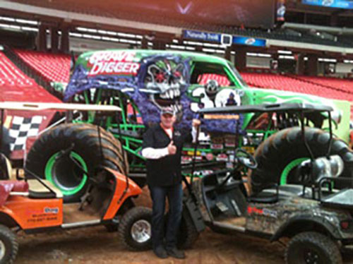 A person stands giving a thumbs up in front of the Grave Digger monster truck, with two golf carts and large tires visible around them inside an arena.