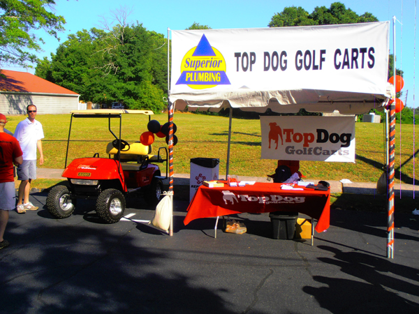 Outdoor booth for Top Dog Golf Carts with a red golf cart, promotional table, and banner; Superior Plumbing sign above. A man stands nearby on a sunny day.