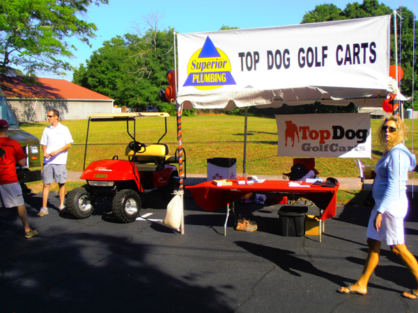 A booth with a "Top Dog Golf Carts" sign, a red golf cart, tables with papers, and people standing and walking nearby on a sunny day.