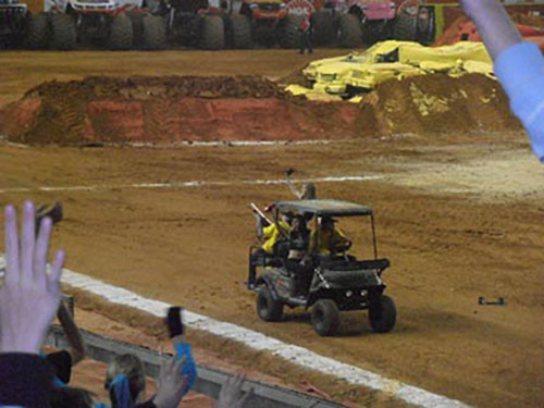 A utility cart carrying several people drives across a dirt arena, with monster trucks and a pile of yellow materials in the background. Spectators in the foreground raise their hands.