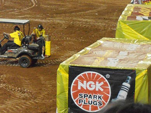 Two workers in yellow shirts sit in a utility vehicle on a dirt arena next to a barricade covered with an NGK Spark Plugs advertisement.