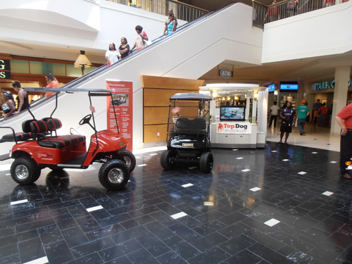 Two golf carts are displayed on a black tiled floor inside a mall near an escalator, with people walking and a kiosk labeled "Top Dog" in the background.