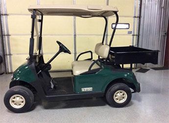 A green golf cart with beige seats and a rear utility cargo bed is parked indoors on a concrete floor.