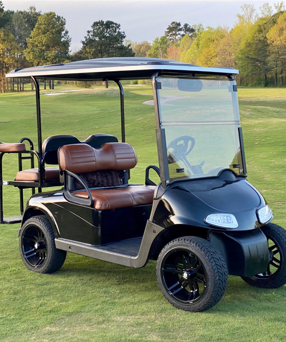 A black golf cart with brown seats is parked on a grassy golf course with trees in the background.