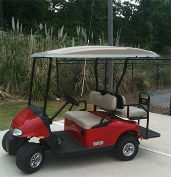 A red golf cart with a roof and two beige bench seats is parked on a concrete surface near a chain-link fence and tall grass.