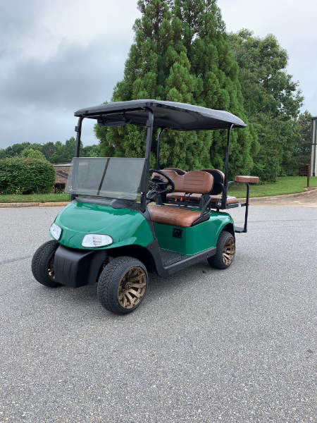 A green golf cart with brown seats is parked on a paved surface near trees and shrubs under a cloudy sky.