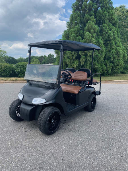 A black golf cart with brown seats is parked on a paved surface, with trees and cloudy sky in the background.