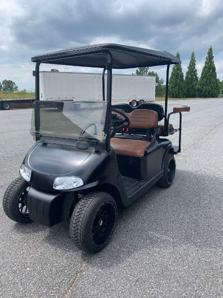 A black golf cart with brown seats is parked on an asphalt surface under a cloudy sky, with trees and a trailer in the background.