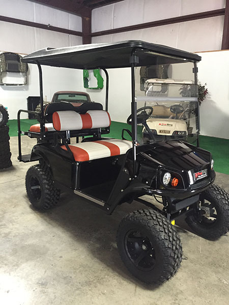 A black golf cart with off-road tires and red-and-white seats is parked indoors on a concrete floor. Another golf cart is visible in the background.