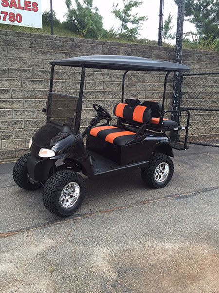 Black golf cart with orange and black seats, parked on pavement near a stone wall and a chain-link fence.
