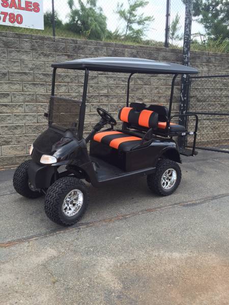 A black golf cart with orange and black seats is parked on pavement near a stone wall and metal fence.