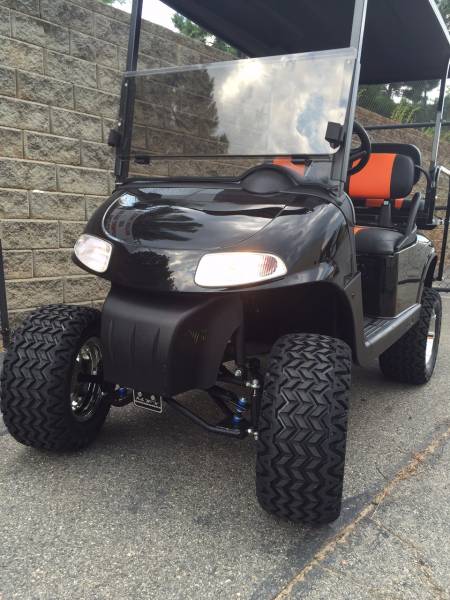 A black golf cart with large off-road tires and orange seats is parked on a paved surface next to a stone wall.