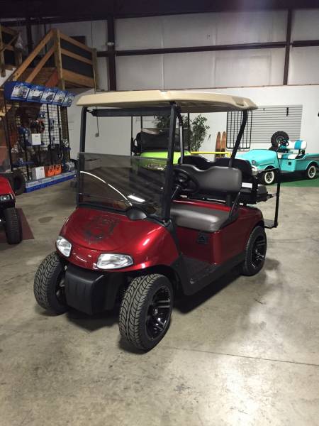 A red golf cart with a beige roof parked indoors on a concrete floor, surrounded by other golf carts and equipment.