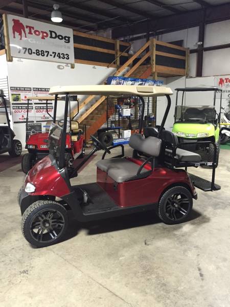 A red golf cart with black wheels is parked inside a showroom with other golf carts, wooden stairs, and signs visible in the background.