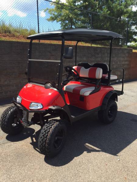 A red and black golf cart with lifted tires and striped seats is parked on pavement near a brick wall and chain-link fence.
