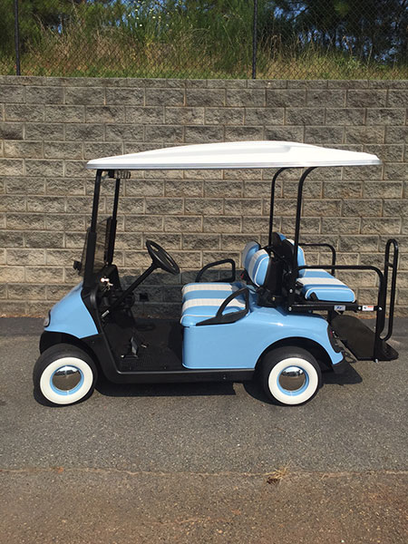 A light blue and white golf cart with two seats, parked on a paved surface in front of a stone wall.