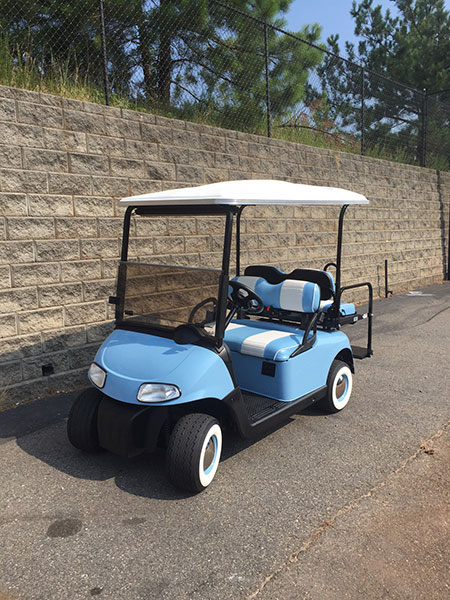 A blue and white golf cart is parked on a paved surface next to a stone wall with trees in the background.