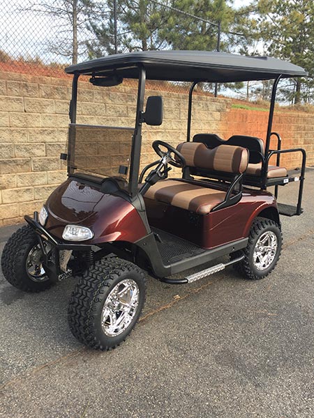 A maroon golf cart with off-road tires and brown cushioned seats is parked on a pavement near a stone wall and chain-link fence.