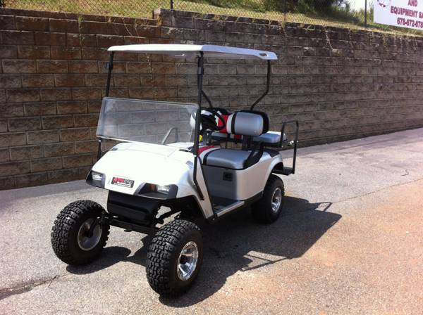 A white golf cart with off-road tires is parked on pavement beside a tall stone retaining wall under a sunny sky.