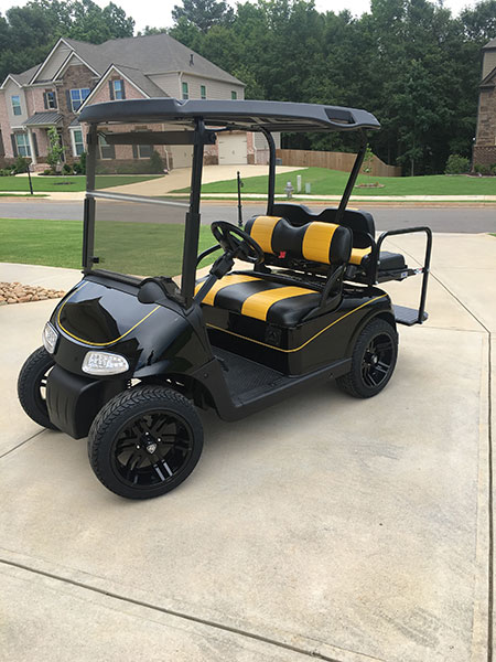A black and yellow golf cart with striped seats is parked on a driveway in a suburban neighborhood.