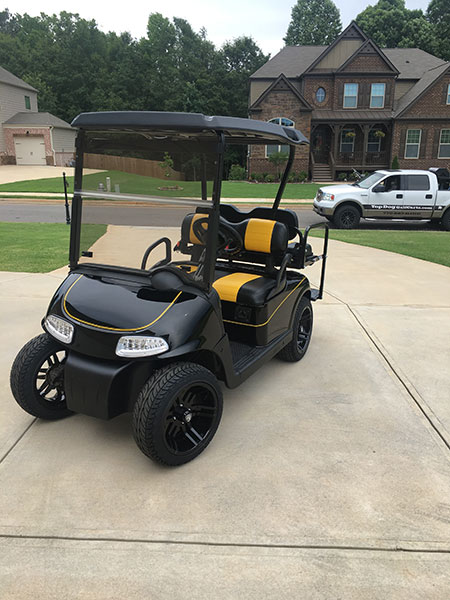A black and yellow golf cart is parked on a concrete driveway in a residential neighborhood, with houses and a white pickup truck in the background.