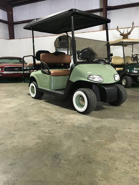 A green golf cart with brown seats and white-rimmed tires is parked indoors on a concrete floor, with vintage cars and a deer mount in the background.