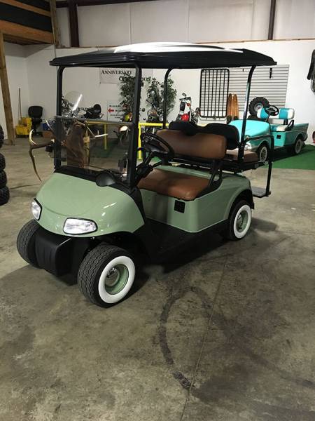 A green golf cart with brown seats and whitewall tires is parked indoors on a concrete floor. Another golf cart and equipment are visible in the background.