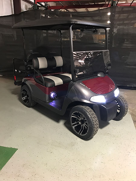 A maroon and black golf cart with two-tone seats and LED headlights is parked indoors on a concrete floor.
