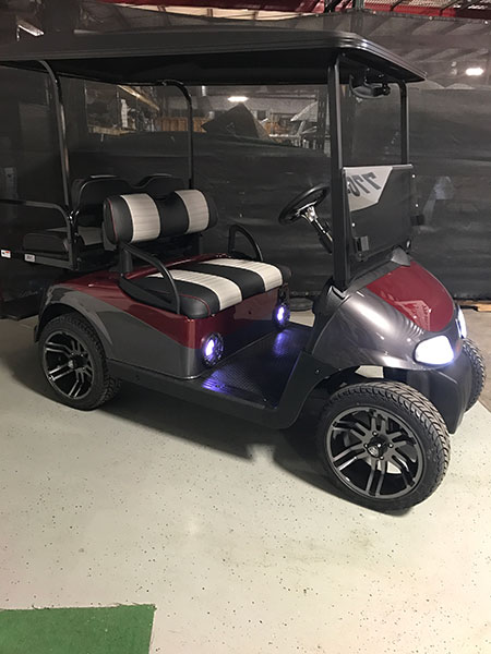 A custom golf cart with black and maroon body, black and silver seats, large alloy wheels, and front headlights parked indoors.