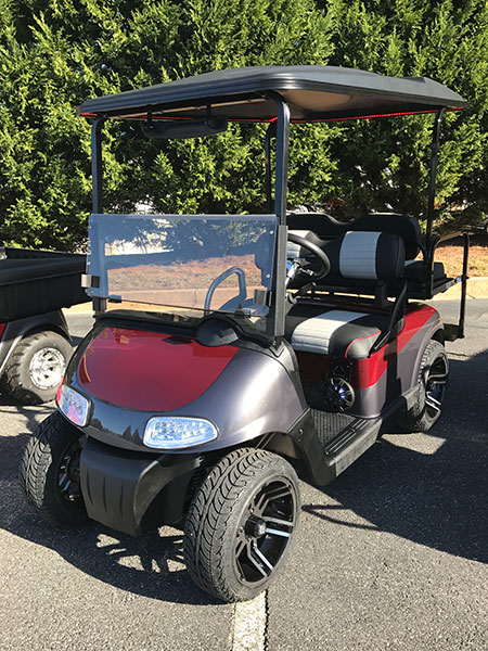 A red and black golf cart with large tires is parked on asphalt next to another cart, with trees in the background.