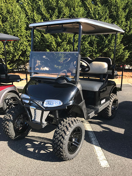A black golf cart with large off-road tires is parked in a parking lot near green trees.
