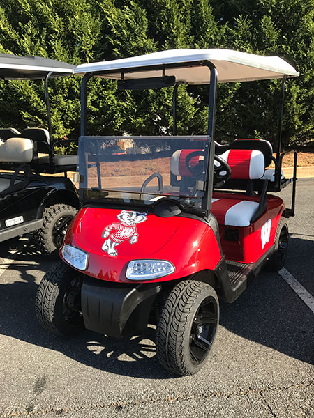 A red golf cart with a white roof and black wheels, featuring a mascot logo on the front and red-and-white striped seats, parked outdoors.