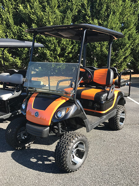 Orange and black golf cart with off-road tires parked on asphalt near green trees.