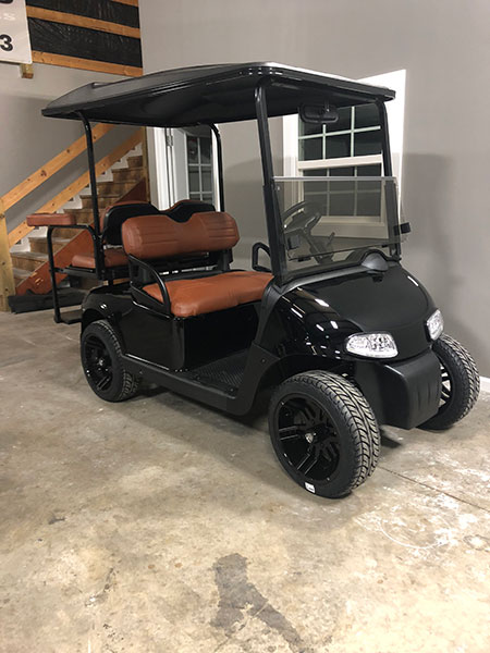 A black golf cart with brown seats is parked indoors on a concrete floor near a staircase and a gray wall with a window.